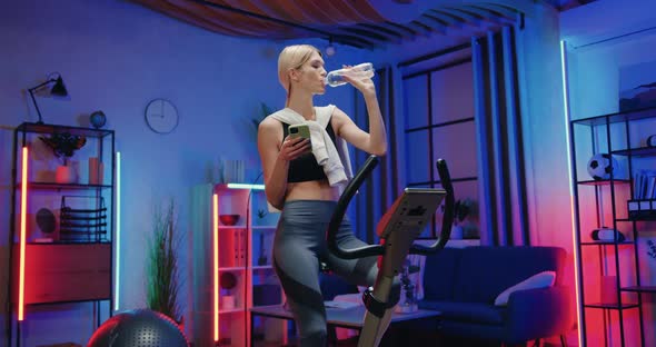 Woman in Sportswear Exercising on Stationary Bike and Drinking Water During Home workout