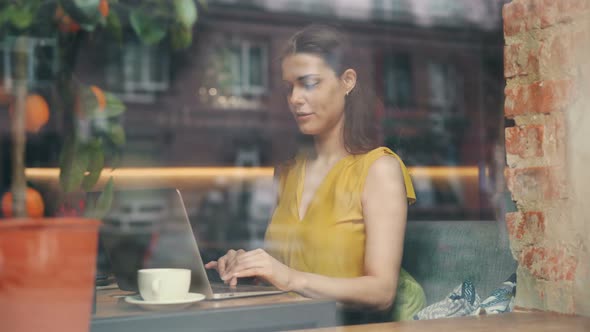 Pretty Lady Using Laptop in Cafe Typing Smiling at Table Behind Glass Window