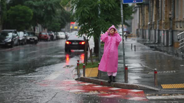 Young Smiling Woman with a Pink Raincoat Crosses the Road on a Rainy Day