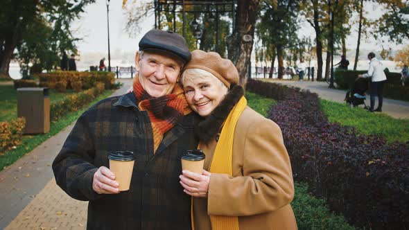 Happy Grandparents Holding Paper Cups of Coffee Smiling and Looking at you Posing at Autumn Park