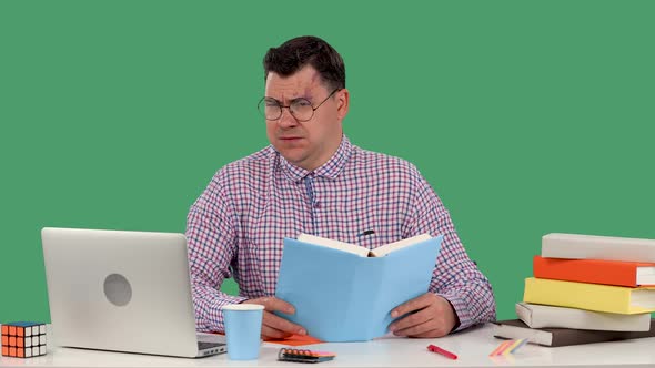 A Man with Glasses Sits at a Desk in Front of a Laptop and Reads a Book Nods His Head Negatively is