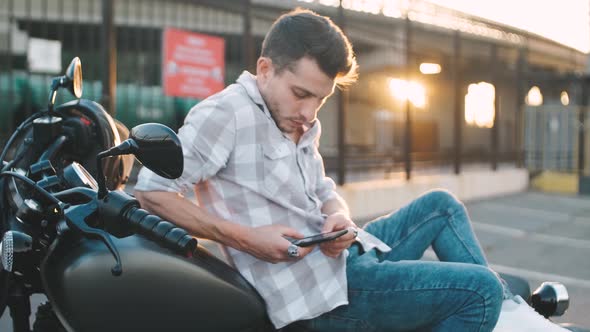 Young Man Motorcyclist with Smart Phone in Hands on Custom Motorcycle on Street