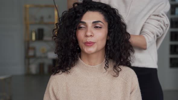 Young Pretty Woman with Curly Black Hair is Sitting in a Chair in a Beauty Salon Her Hair is Being