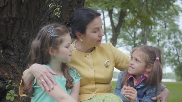 Portrait Elegant Senior Grandmother Sitting on the Grass Under the Tree in the Park Hugging Granddaughters