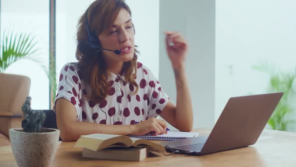 Female Call Center Worker in Headset Looks at Laptop Screen Write in Notebook