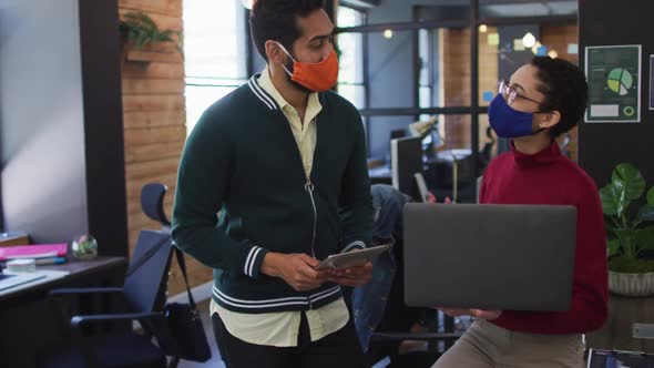 Diverse male and female colleagues wearing face masks using laptop digital tablet discussing at mode