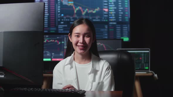 Asian Female Stock Market Broker Working On Computer And Smiling To Camera With Analyzing Graphs