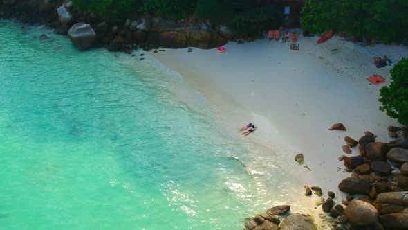 An aerial shot of white sand beach with large stones, clear blue water and unrecognisable people on