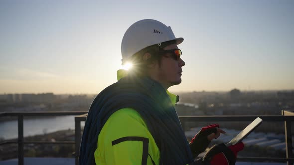 Industrial Climber in a White Helmet Walks Along the Roof with a Tablet and a Rope Against the