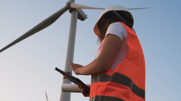 Female an Engineer in a Helmet and an Orange Vest Controls the Operation of a Wind Turbine Using a