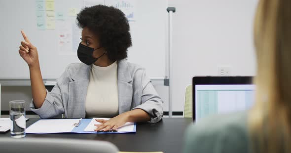 African american businesswoman wearing face mask sitting at desk talking to colleagues in office