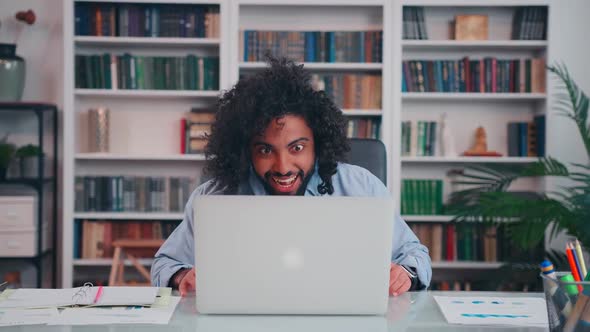 Overjoyed Young Arabian Man Yelling and Waving Hand Sits at Table with Laptop