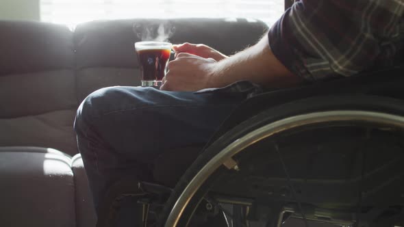 Midsection of caucasian disabled man in wheelchair holding cup of coffee in living room