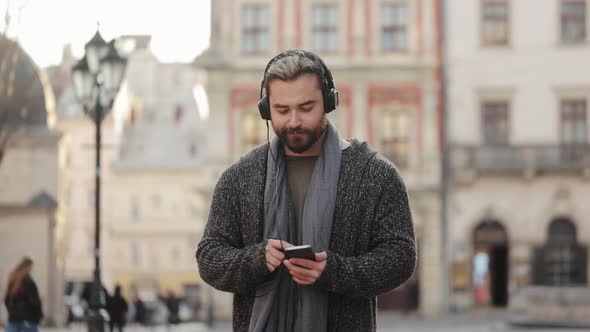 A Happy Bearded Man is Listening to Music with Headphones and Scrolling Through Social Networks on