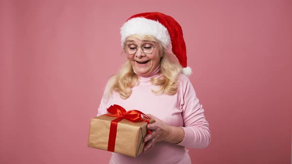 Happy Curious Senior Woman in Santa Hat Shaking Gift Box and Surprising Pink Background Fast Motion