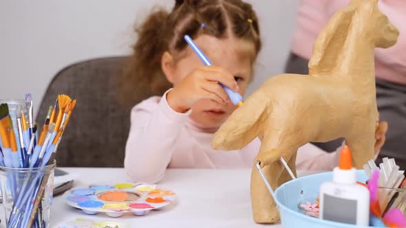 Step by step. Little girl painting paper mache unicorn with blue paint.