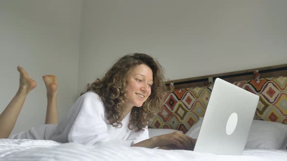 Happy Woman Falls on Bed Opens Laptop Smiling and Types
