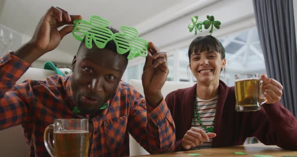 Portrait of happy diverse couple with beer wearing clover shape items and having video call