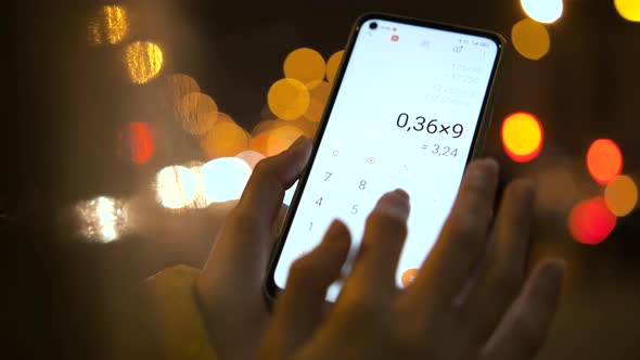 Female hands holding smartphone using calculator app to count nubmers outside at night.