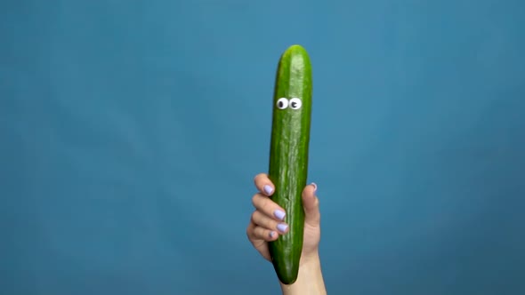 Cucumber with Eyes in a Woman Hand Close-up. Cucumber Shakes and Twists Eyes on a Blue Background