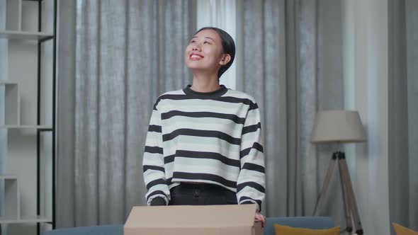 Smiling Young Asian Woman Carrying Cardboard Box With Stuff Into A New House Then Looking Around
