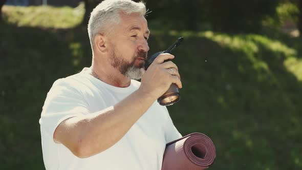 Senior Man Drinking Water and Holding Exercise Mat Outside