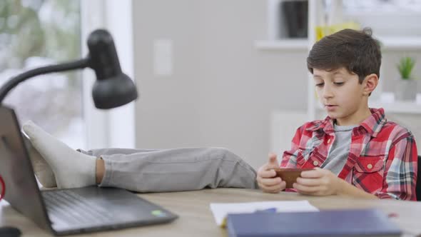 Relaxed Middle Eastern Little Boy Sitting with Feet on Table and Messaging Online on Smartphone