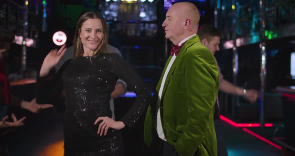 Middle Shot of Young Cheerful Caucasian Woman in Black Dress Dancing in Night Club Next To Elegant