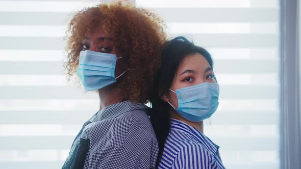 African American and Asian Women with Medical Mask Standing Back To Back in Front of the Window