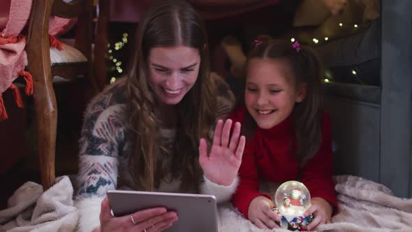 Caucasian mother and daughter waving while having a video chat on digital tablet under blanket fort