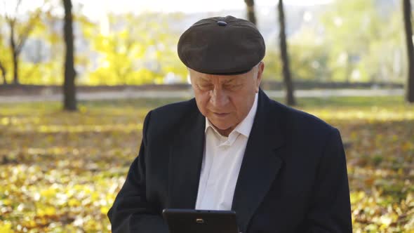 Senior Man in Cap Using Tablet and Telling on Sunny Autumn Background