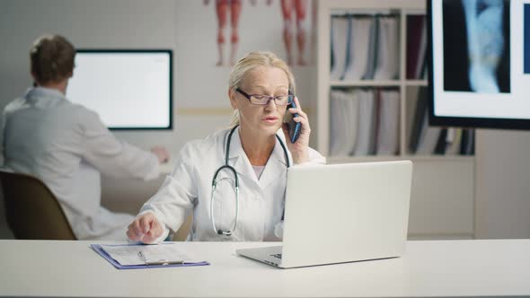 Attractive Mature Doctor Talking on Smartphone in Office Consulting Patient.