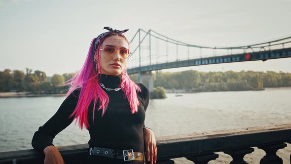 Young Hipster Lady with Pink Hair is Looking Away and Then at you While Posing Leaning on Fence of