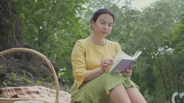 Portrait of an Elegant Beautiful Mature Woman Reading the Book Sitting on the Blanket Under the Tree