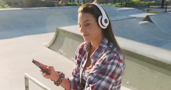 Smiling caucasian woman wearing headphones, using smartphone and waving at a skatepark