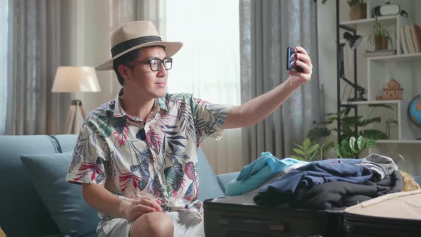 Asian Young Man Try Hat And Selfie While Packing Clothes In Suitcase At Home, Preparing For Vacation