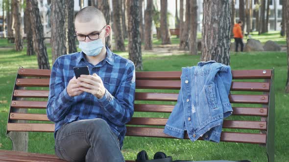 Guy in protective medical mask with smartphone in hands sitting on bench in city park. 