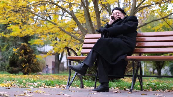 Elderly Grandfather Sitting on a Bench and Talking on the Phone in the Autumn Park