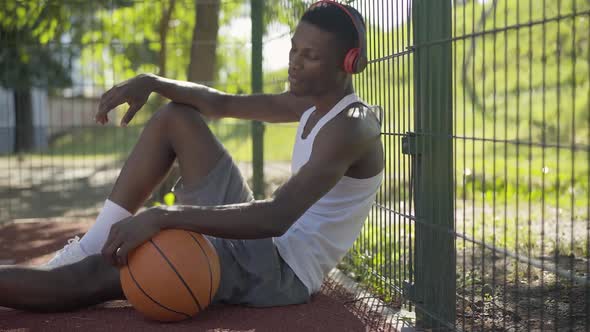 Side View of Positive African American Sportsman Sitting with Ball on Basketball Court and Listening