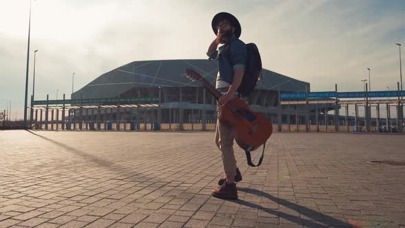 Hipster Bearded Male Musician with Dark Warm Eyes Walking in the Park on a Sunny Day with a Guitar