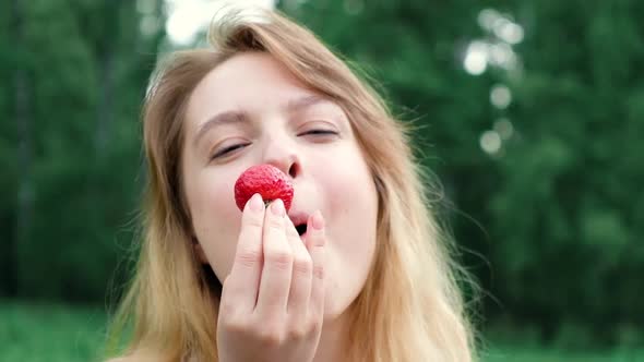 Portrait of girl biting ripe strawberry in the forest on a picnic on summer day