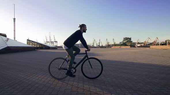 Stylish Handsome Man Riding Bicycle During Sunset or Sunrise with Sea Port on Background