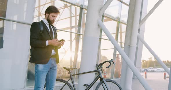 Man Standing Near Glass Wall with His Bike and Using Phone