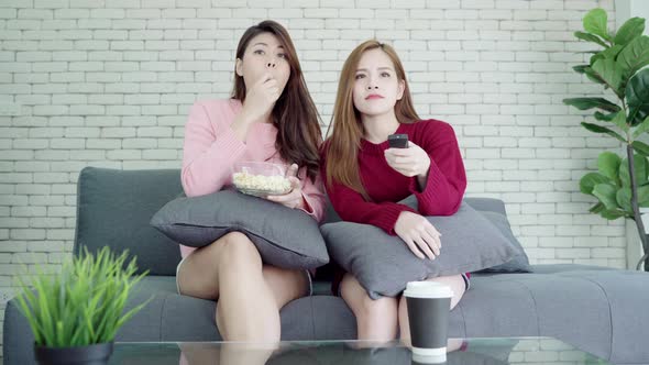 Lesbian Asian couple watching TV laugh and eating popcorn in living room.