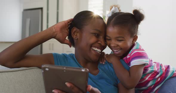 Happy african american mother and daughter sitting on sofa using digital tablet and laughing