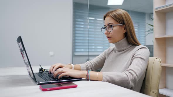 Cute Woman Working at a Laptop with Glasses Typing Text in the Office Employee at Work on the