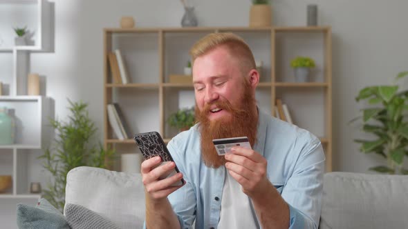 Redhead Beard Man Sit on Couch Holding Credit Card and Cellphone Make Easy Remote Electronic
