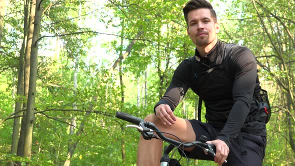 A Young Handsome Cyclist Sits on His Bike in a Forest and Talks To the Camera - View From Below