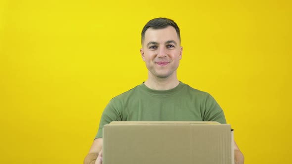 Young Delivery Man Courier in a Green Tshirt Lifts a Craft Cardboard Box in Front of Him