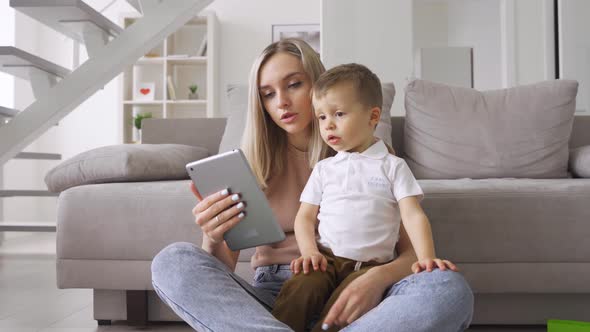 Young Mom Sitting on Floor at Home with Kid Watching Movies on Tablet